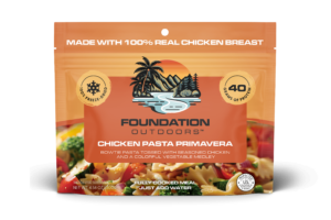 Picture of the front of an orange product pouch containing freeze dried Chicken Pasta Primavera Meal. The logo contains green palm trees on the left, green pine trees on the right, brown mountains in the background, blue water running the middle, and an orange setting sun the background. Below the logo is a picture of the meal which show bowtie pasta, green broccoli, red tomatoes, and yellow zucchini half moon slices.